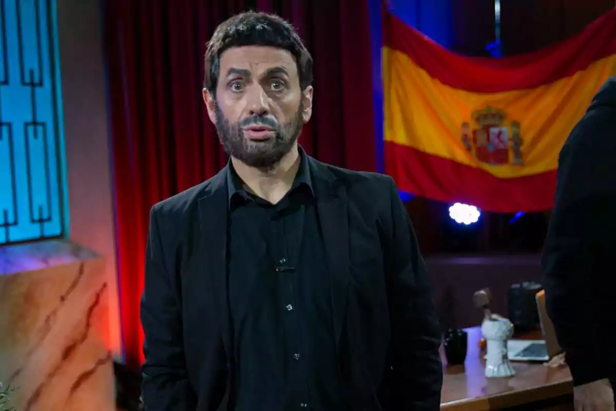 José Mota amb barba i vestit fosc està dret davant d'una bandera d'Espanya en un escenari il·luminat. José Mota amb barba i vestit fosc està dret davant d'una bandera d'Espanya en un escenari il·luminat.