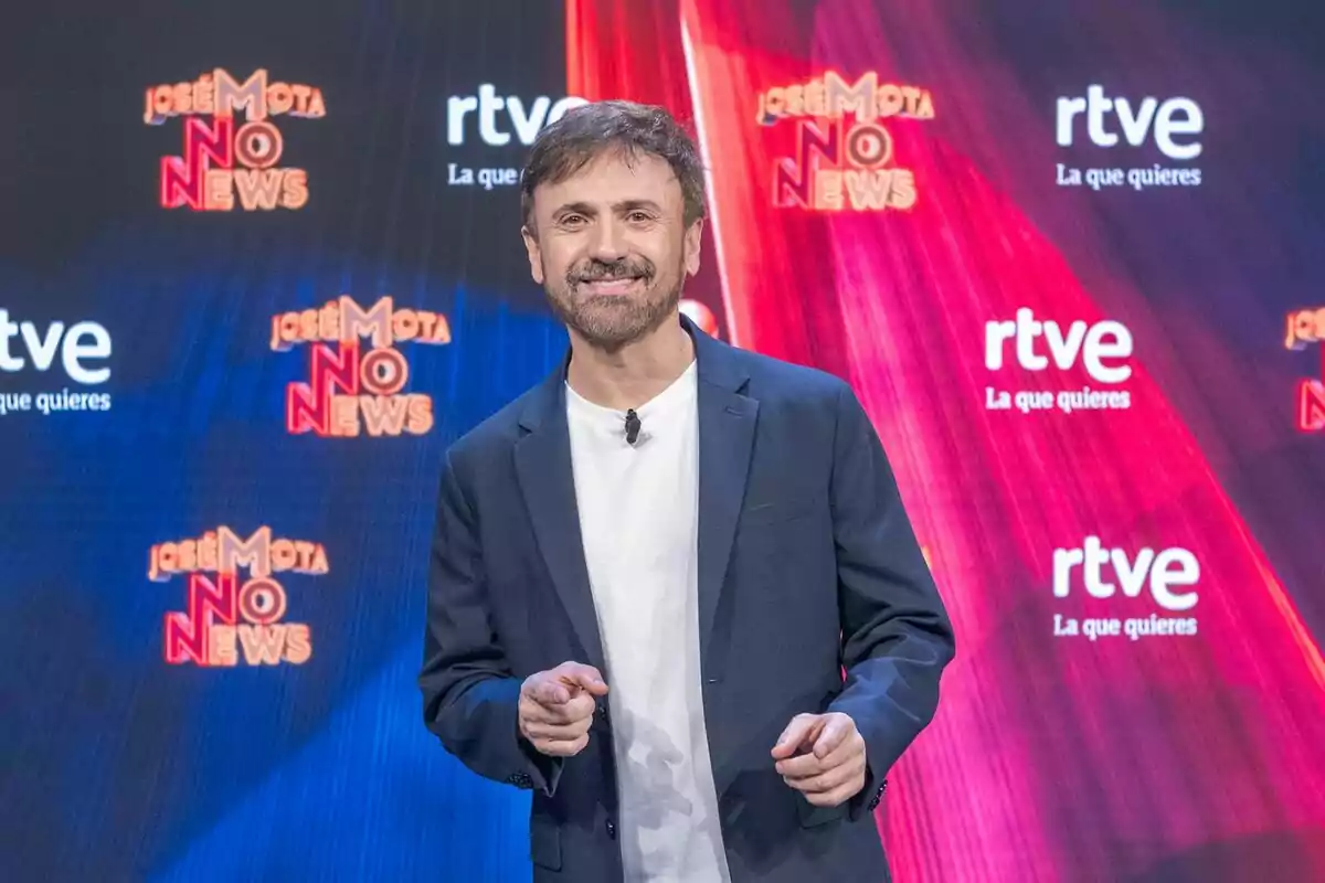 José Mota sonriente con barba y cabello corto, vestido con camiseta blanca y chaqueta oscura, está de pie frente a un fondo con logotipos de José Mota sonriente con barba y cabello corto, vestido con camiseta blanca y chaqueta oscura, está de pie frente a un fondo con logotipos de