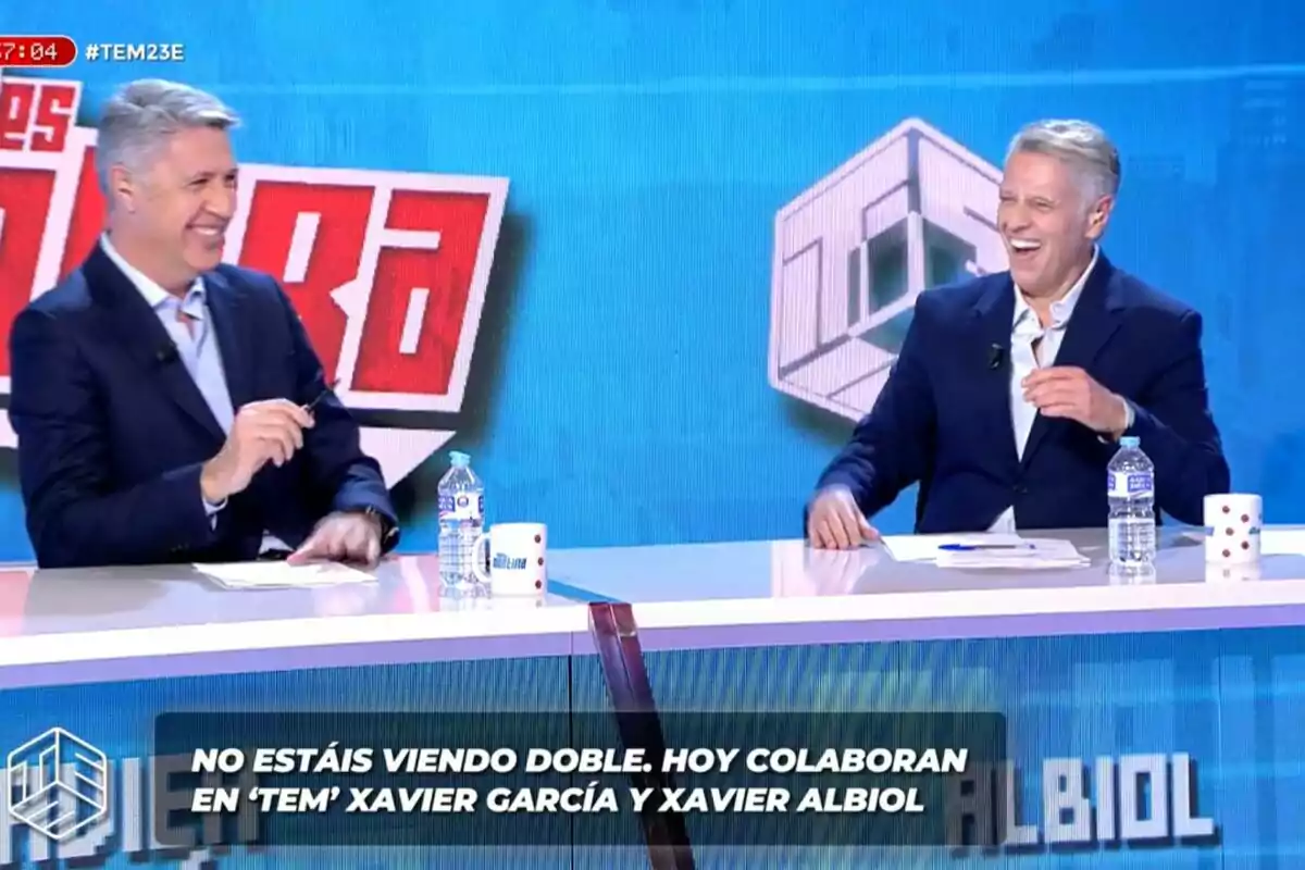 Josep Ferré imitando a Xavier Albiol sonrientes mientras están sentados en un set de televisión con tazas y botellas de agua sobre la mesa, y un texto en la parte inferior menciona que no se está viendo doble. Josep Ferré imitando a Xavier Albiol sonrientes mientras están sentados en un set de televisión con tazas y botellas de agua sobre la mesa, y un texto en la parte inferior menciona que no se está viendo doble.