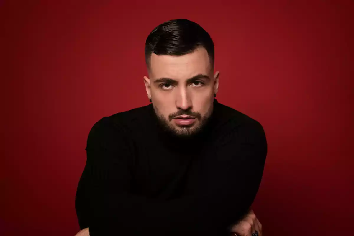 Joshua Velázquez con barba y cabello corto viste un suéter negro posando frente a un fondo rojo. Joshua Velázquez con barba y cabello corto viste un suéter negro posando frente a un fondo rojo.