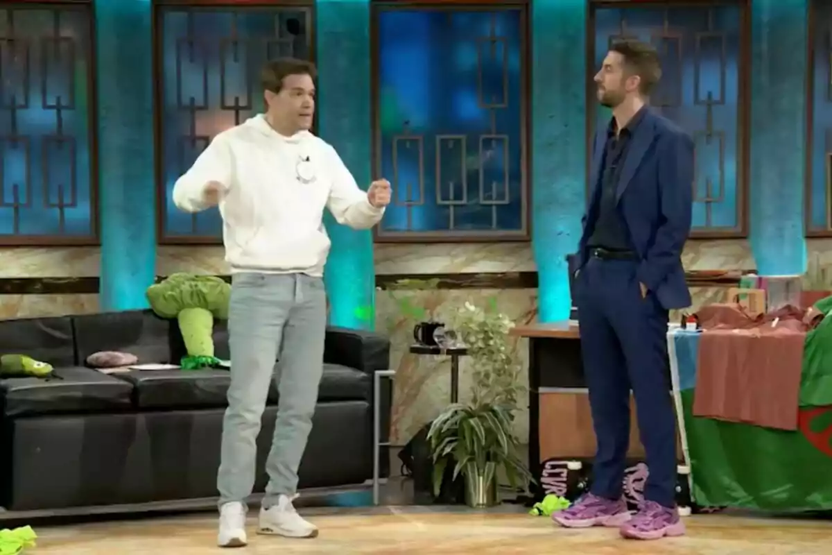 Juan Dávila y David Broncano están de pie en 'La Revuelta'; una lleva sudadera blanca y jeans, mientras que la otra viste un traje azul y zapatillas rosas grandes; el fondo tiene un sofá negro con cojines y decoraciones coloridas. Juan Dávila y David Broncano están de pie en 'La Revuelta'; una lleva sudadera blanca y jeans, mientras que la otra viste un traje azul y zapatillas rosas grandes; el fondo tiene un sofá negro con cojines y decoraciones coloridas.