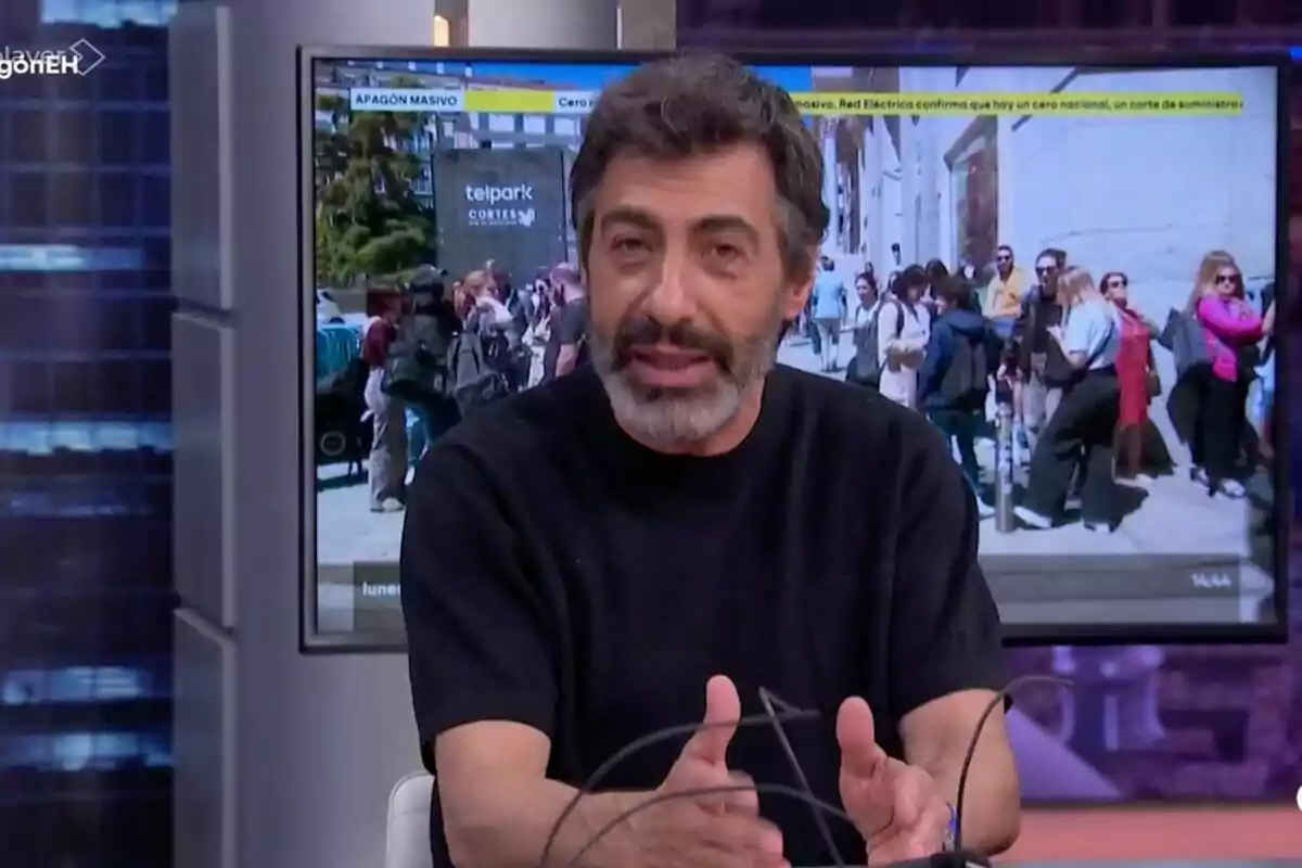 Juan del Val con barba y cabello oscuro está hablando en un programa de televisión con una pantalla de fondo que muestra una multitud de personas en la calle.