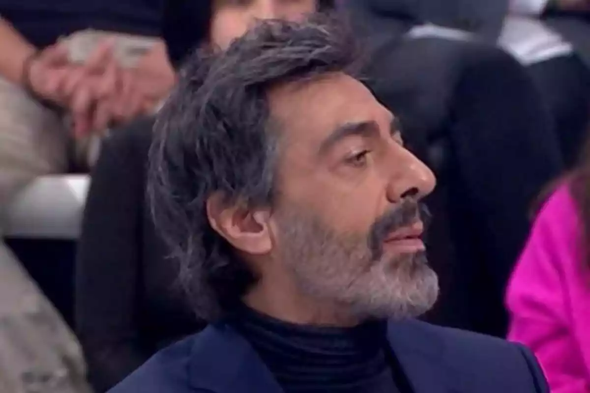 Plano corto de Juan del Val de perfil con barba y cabello oscuro en un entorno de interior con personas al fondo. Plano corto de Juan del Val de perfil con barba y cabello oscuro en un entorno de interior con personas al fondo.
