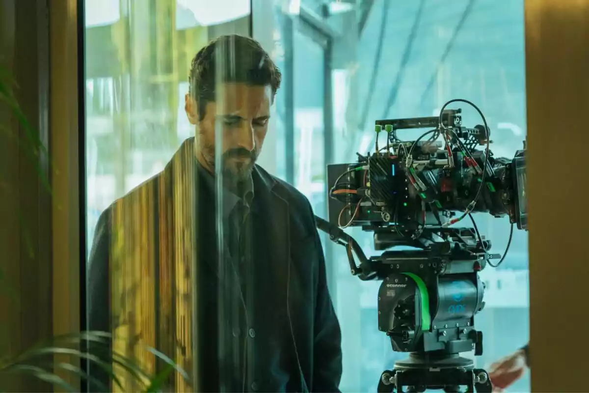Juan Diego Botto de pie junto a una cámara de cine en un entorno moderno con reflejos en el vidrio en el rodaje de El Centro. Juan Diego Botto de pie junto a una cámara de cine en un entorno moderno con reflejos en el vidrio en el rodaje de El Centro.