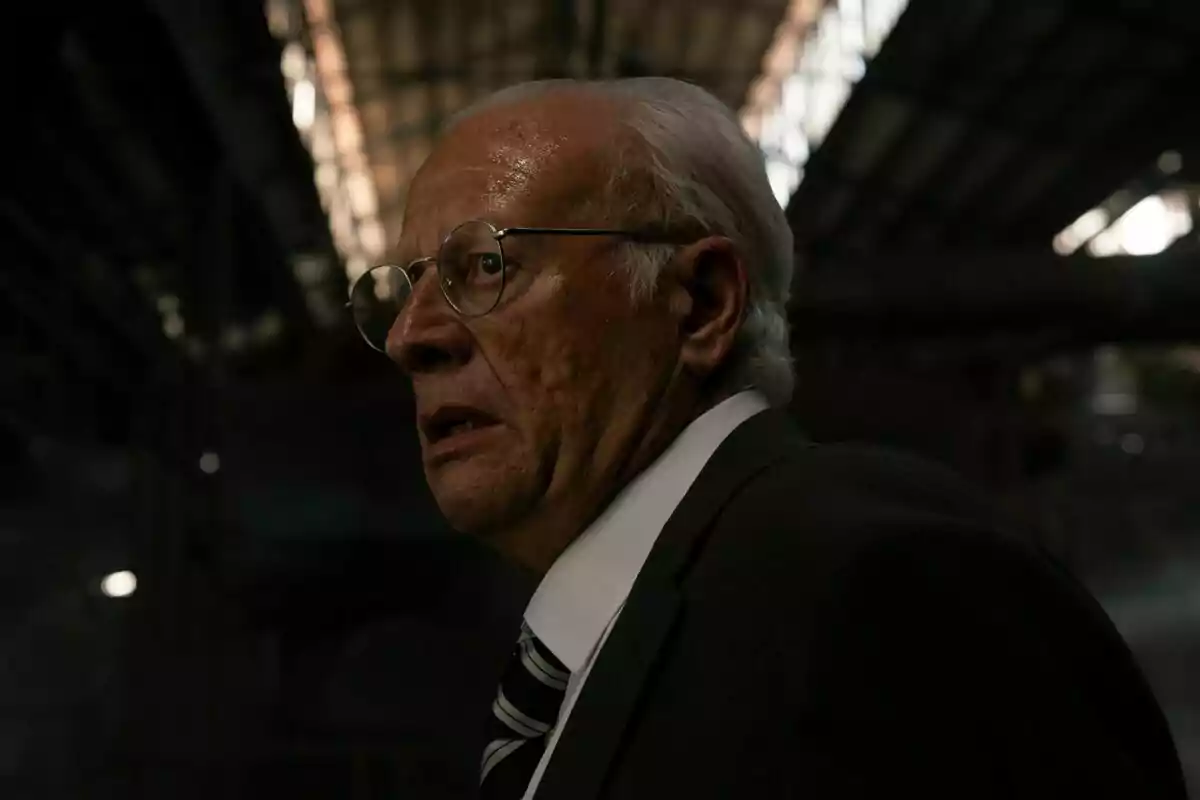 Juan Echanove mayor con gafas y traje oscuro está en un ambiente industrial con poca luz en Memento Mori.