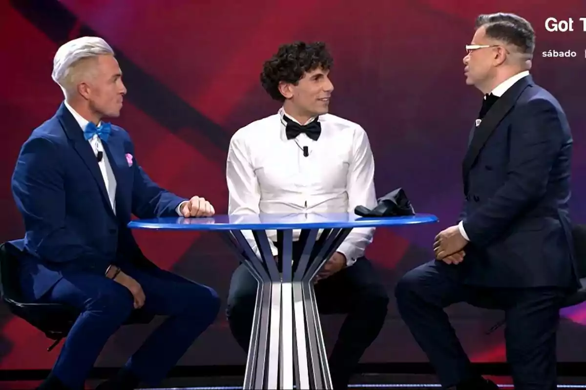 Juan, Óscar y Jorge Javier Vázquez vestidos formalmente conversan alrededor de una mesa azul en Gran Hermano con fondo rojo. Juan, Óscar y Jorge Javier Vázquez vestidos formalmente conversan alrededor de una mesa azul en Gran Hermano con fondo rojo.