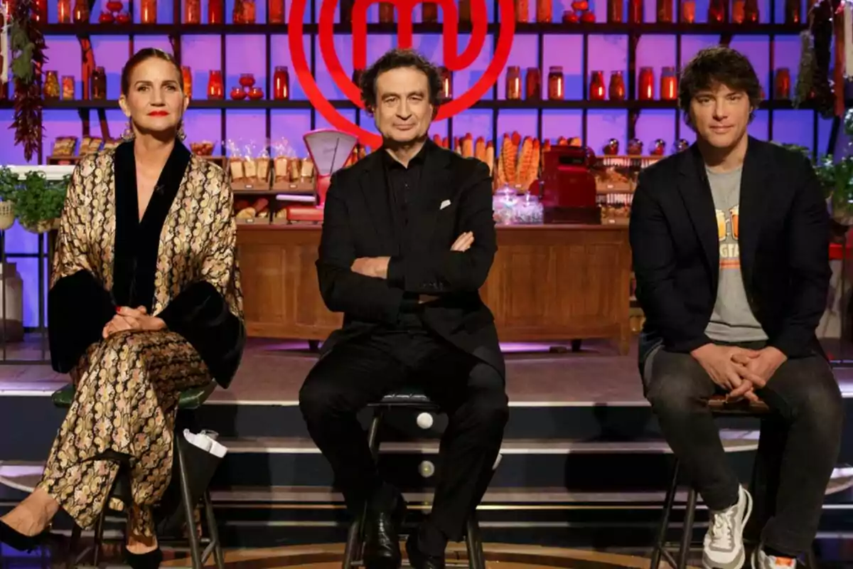 Pepe Rodríguez, Samantha Vallejo-Nágera y Jordi Cruz están sentadas en taburetes frente a un fondo de cocina iluminado con luces moradas y decorado con estantes llenos de ingredientes y utensilios culinarios en MasterChef.