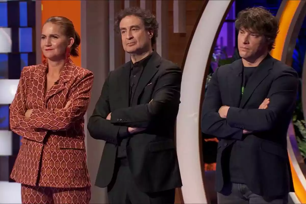 Samantha Vallejo-Nágera, Pepe Rodríguez y Jordi Cruz de pie con los brazos cruzados, vestidas de manera formal, en un entorno iluminado y colorido en MasterChef.