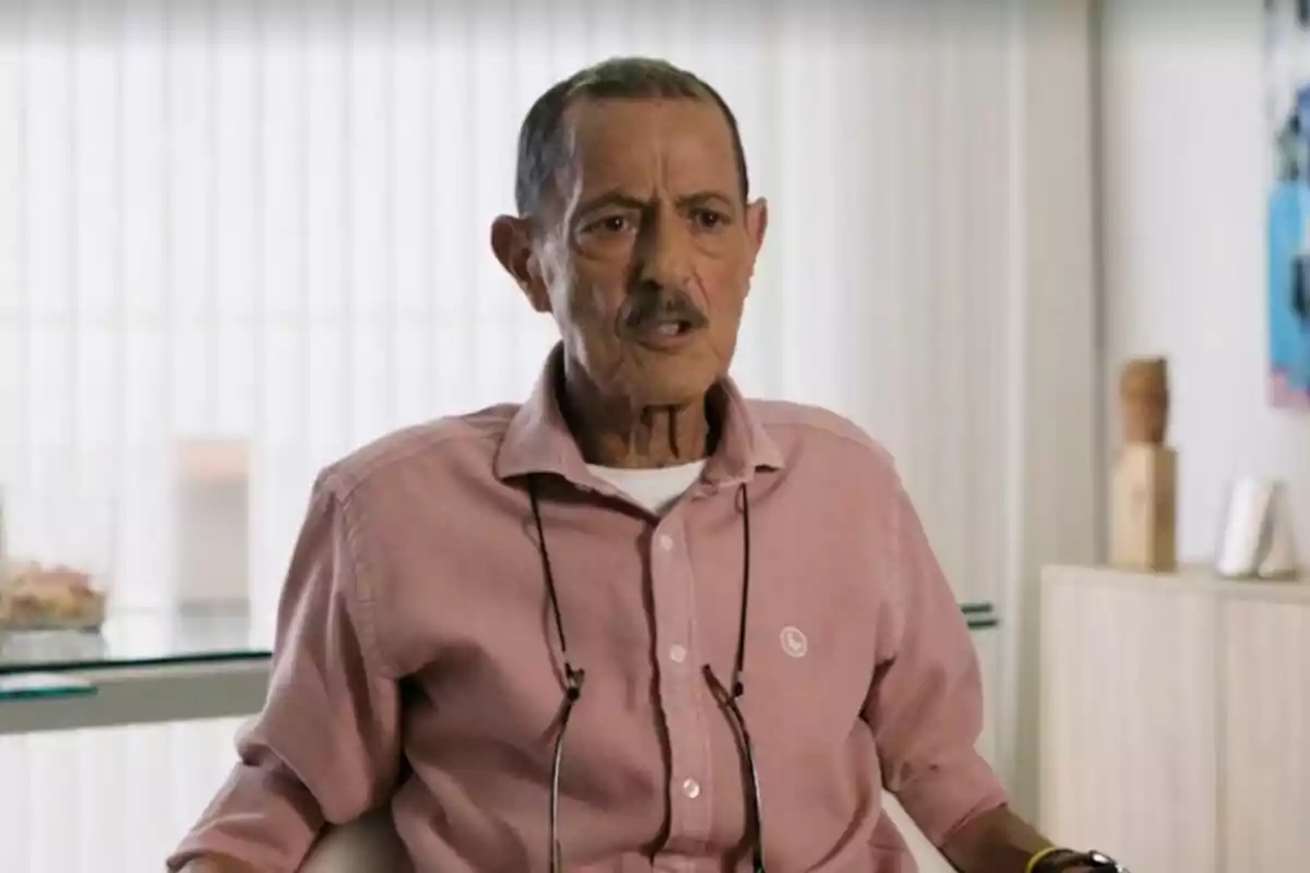 Julián Muñoz con bigote y camisa rosa está sentado en una silla en una habitación con cortinas blancas y muebles claros. Julián Muñoz con bigote y camisa rosa está sentado en una silla en una habitación con cortinas blancas y muebles claros.