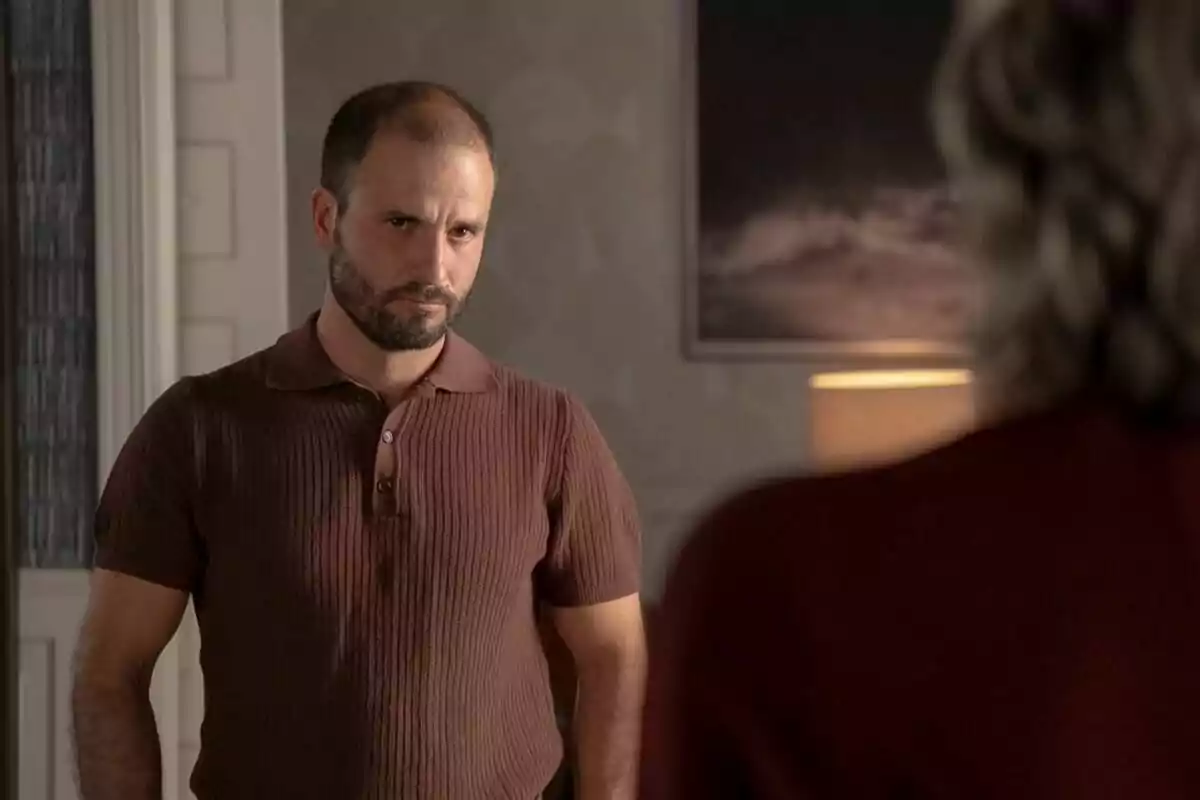 Julián Ortega con barba y camiseta marrón está mirando seriamente a otra persona en una habitación iluminada con una lámpara y un cuadro en la pared en la serie 4 estrellas. Julián Ortega con barba y camiseta marrón está mirando seriamente a otra persona en una habitación iluminada con una lámpara y un cuadro en la pared en la serie 4 estrellas.