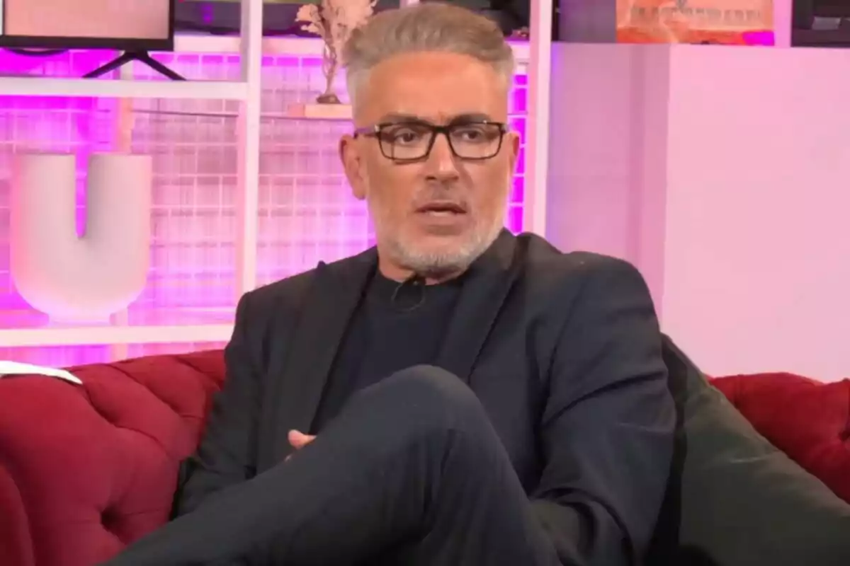 Kiko Hernández con gafas y cabello canoso está sentado en un sofá rojo en un ambiente iluminado con luces rosadas en 'Tentáculos'.
