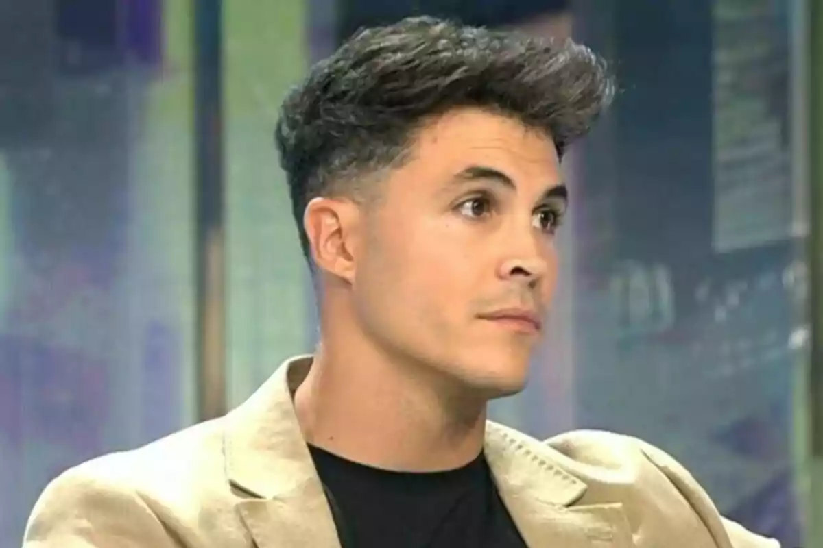 Kiko Jiménez con cabello corto y oscuro, vestido con una chaqueta beige y una camiseta negra, mira hacia un lado con una expresión seria en su rostro en De Viernes. Kiko Jiménez con cabello corto y oscuro, vestido con una chaqueta beige y una camiseta negra, mira hacia un lado con una expresión seria en su rostro en De Viernes.