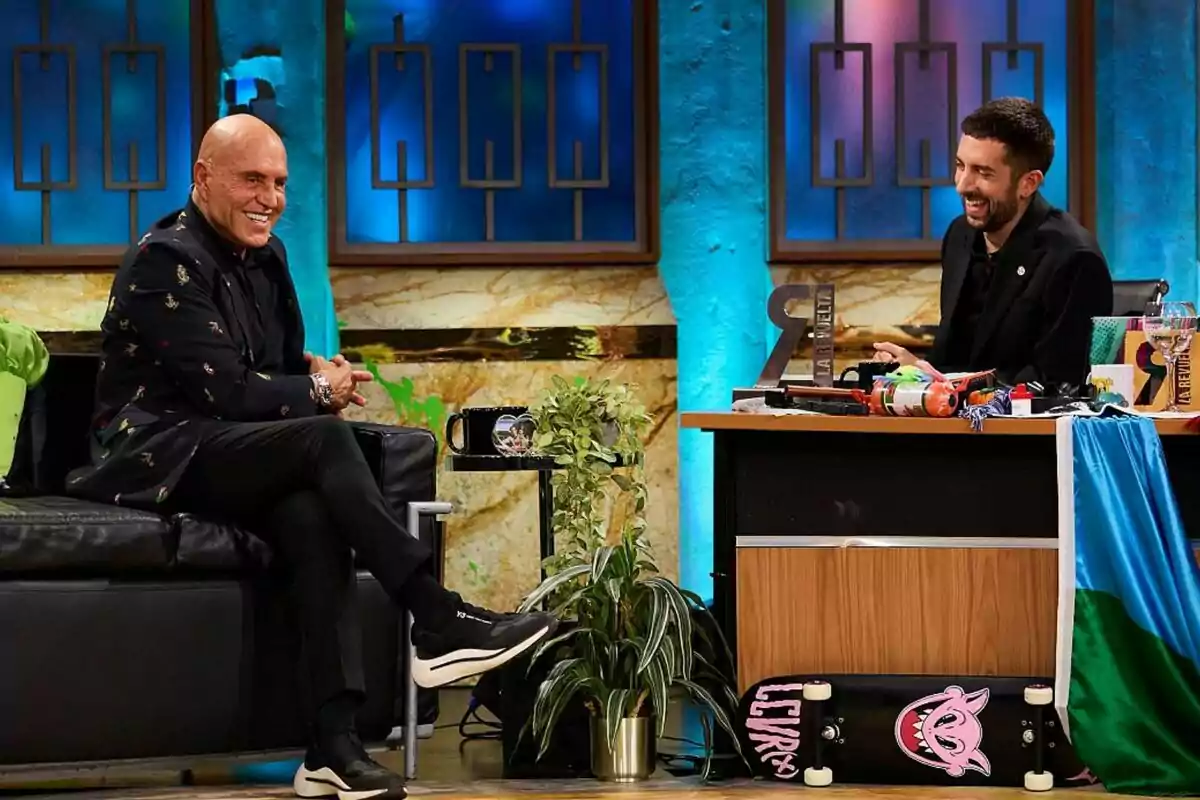 Kiko Matamoros amb jaqueta fosca i sabatilles esportives està assegut en un sofà negre, somrient i conversant amb David Broncano que està darrere d'un escriptori ple d'objectes colorits, a La Revuelta amb llums blaves i verdes. Kiko Matamoros amb jaqueta fosca i sabatilles esportives està assegut en un sofà negre, somrient i conversant amb David Broncano que està darrere d'un escriptori ple d'objectes colorits, a La Revuelta amb llums blaves i verdes.