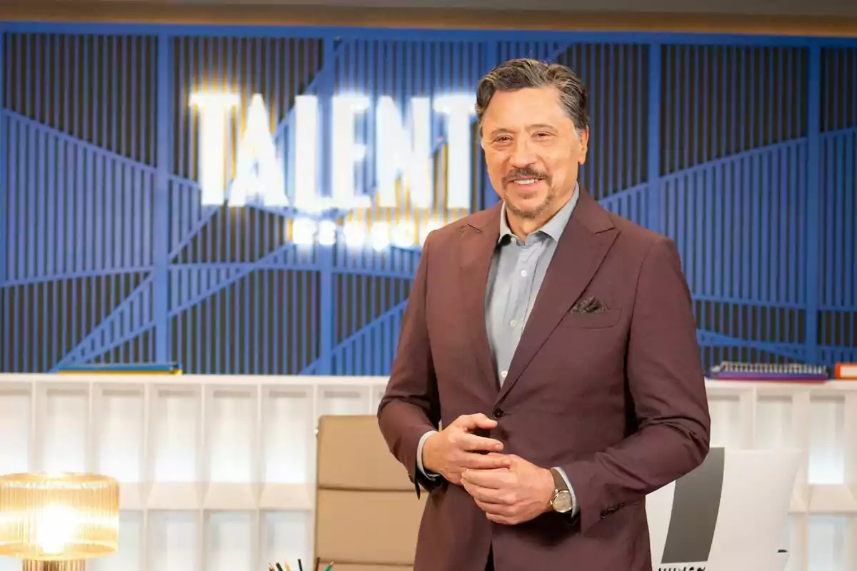 Carlos Bardem amb vestit marró fosc i camisa blau clar està dret davant d'un fons blau amb la paraula 