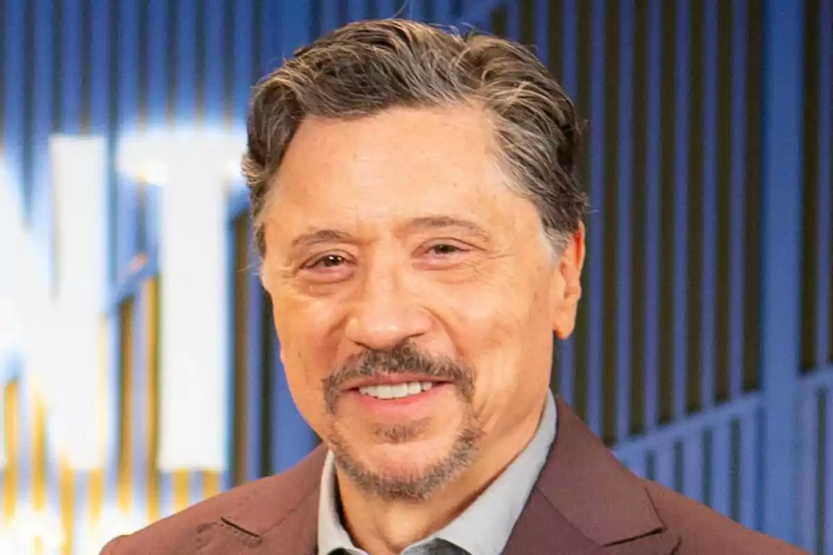 Carlos Bardem amb cabell fosc i barba somriu davant d'un fons blau amb línies verticals a La Agència.