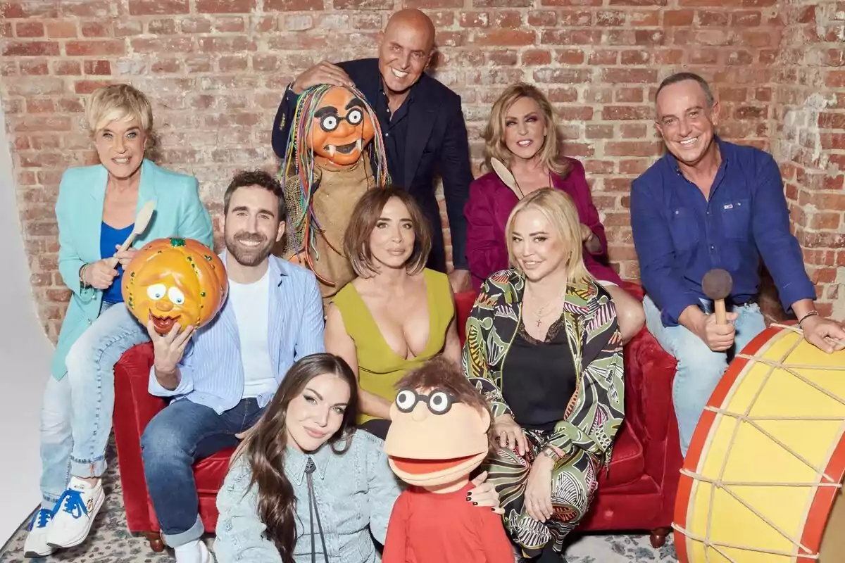 Un grup de persones format per María Patiño, Aitor Albizua, Belén Esteban, Inés Hernand, Chelo García Cortés, Kiko Matamoros, Lydia Lozano i Víctor Sandoval posant en un sofà vermell amb titelles i un tambor gran, tots somriuen davant d'una paret de maons a La Família de la Tele.