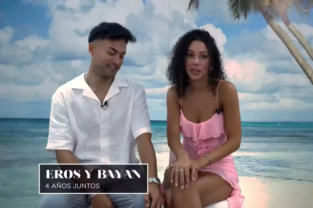 Eros i Bayan asseguts davant d'un fons de platja amb palmeres i cel blau. Captura de 'La Isla de las Tentaciones'.