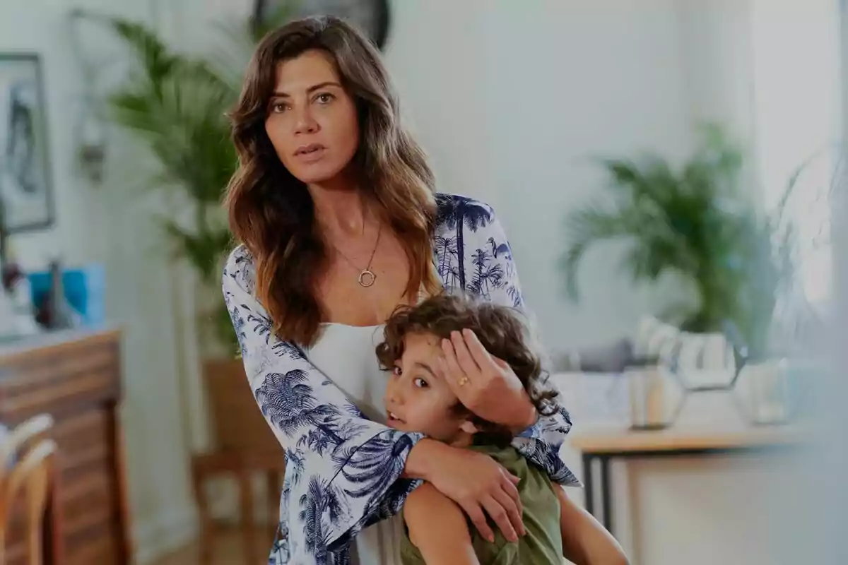 Una mujer con cabello largo y ondulado abraza a un niño pequeño en una habitación iluminada y decorada con plantas en la serie La ley del amor Una mujer con cabello largo y ondulado abraza a un niño pequeño en una habitación iluminada y decorada con plantas en la serie La ley del amor
