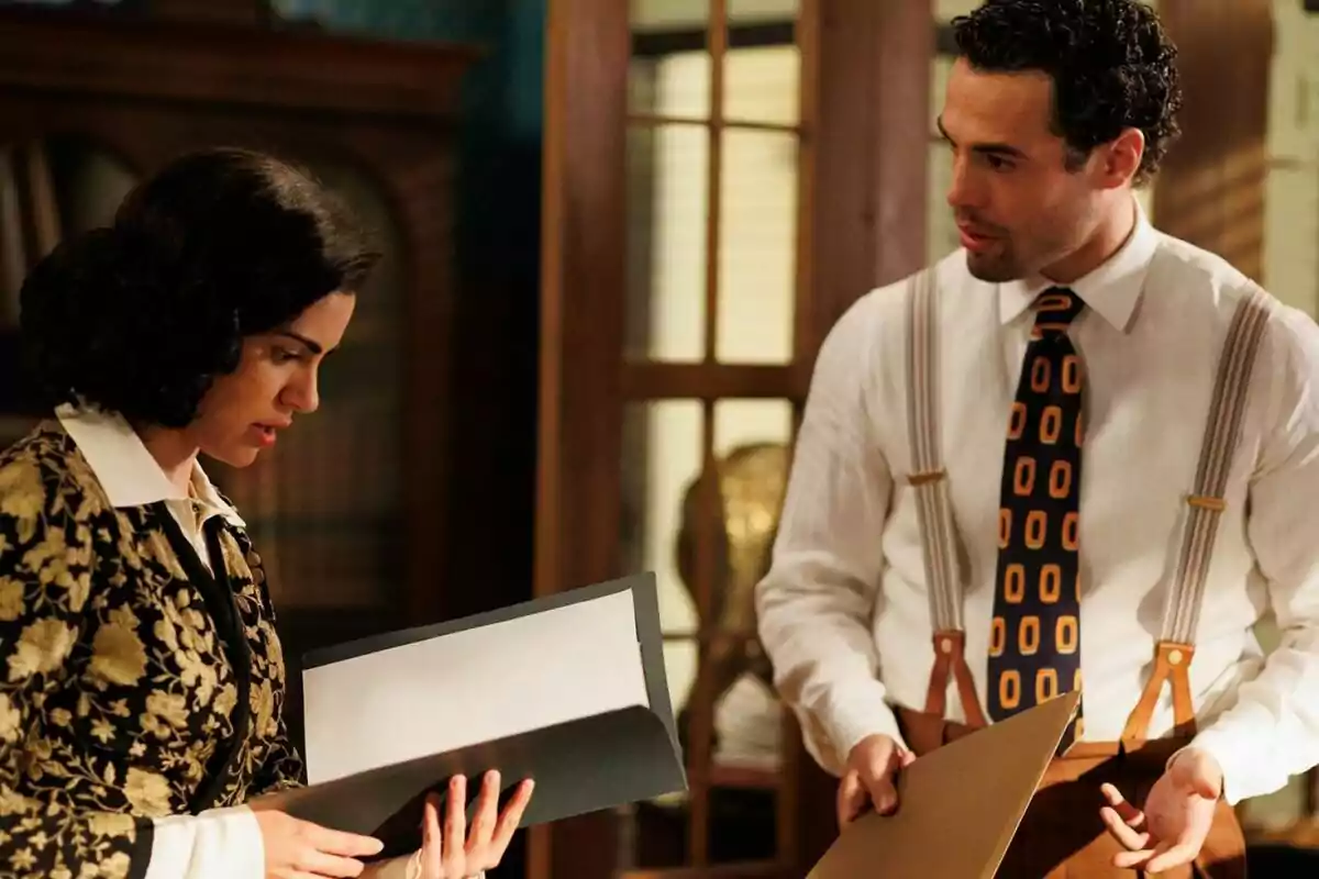 Paula y Rodrigo vestidas con ropa de época están revisando documentos en una oficina con muebles de madera. Captura de 'La Moderna' Paula y Rodrigo vestidas con ropa de época están revisando documentos en una oficina con muebles de madera. Captura de 'La Moderna'