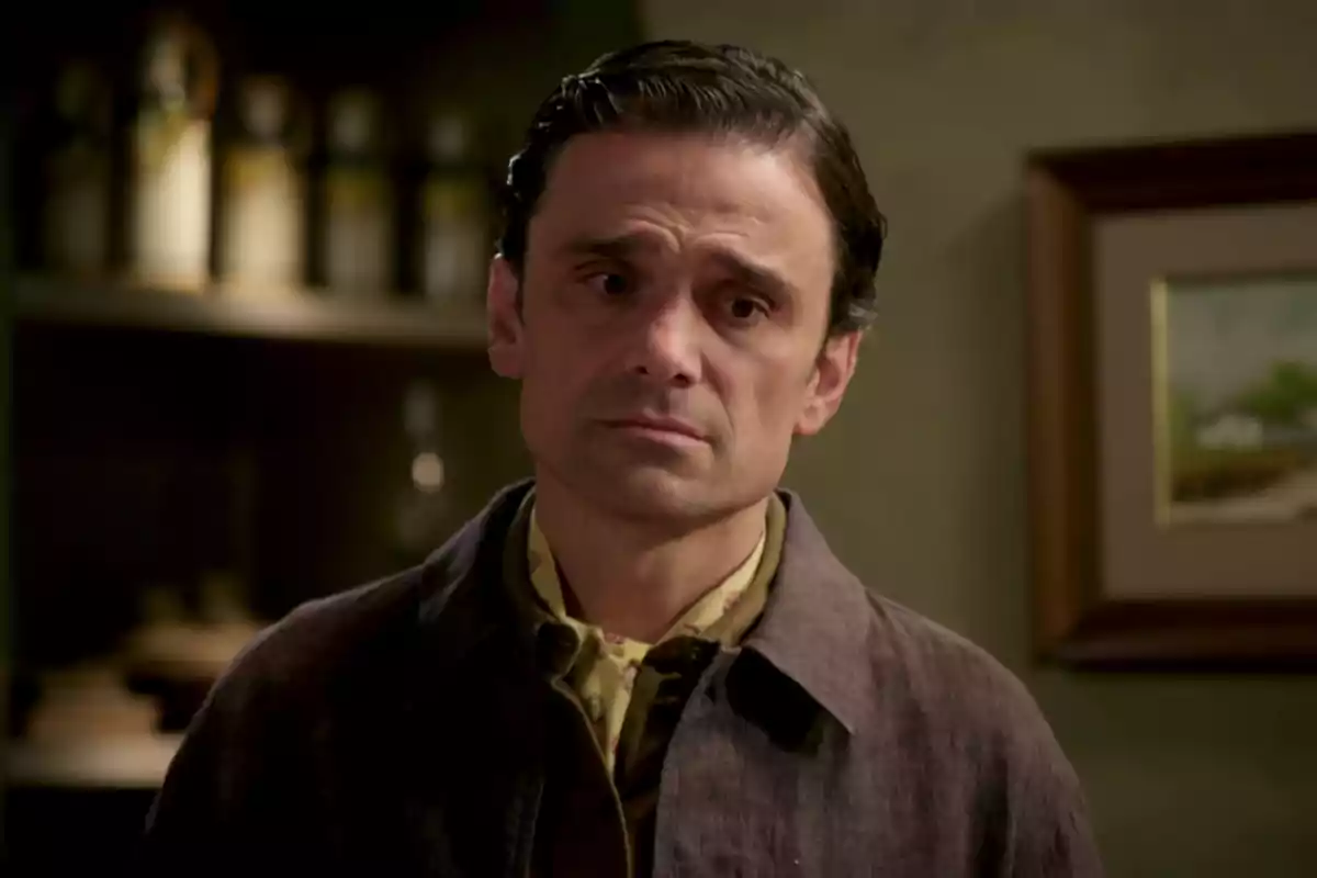 Pietro amb expressió seriosa i cabell fosc està en una habitació amb prestatges i un quadre a la paret.