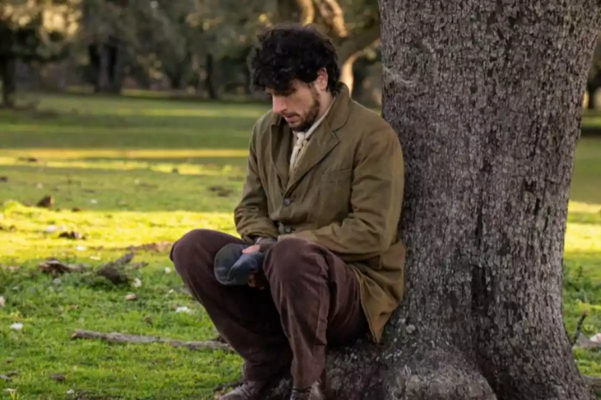 Antonio con chaqueta marrón y pantalones oscuros está sentado pensativo al pie de un árbol en un parque. Captura de 'La Promesa' Antonio con chaqueta marrón y pantalones oscuros está sentado pensativo al pie de un árbol en un parque. Captura de 'La Promesa'