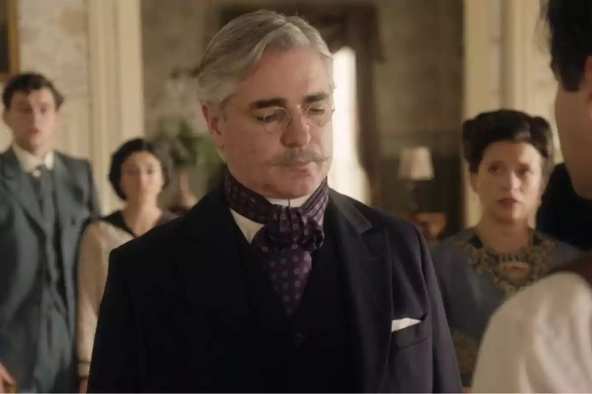El doctor Gamarra con bigote y gafas, vestido elegantemente con un traje oscuro y una corbata de lazo, está en una habitación con otras personas al fondo. Captura de 'La Promesa' El doctor Gamarra con bigote y gafas, vestido elegantemente con un traje oscuro y una corbata de lazo, está en una habitación con otras personas al fondo. Captura de 'La Promesa'