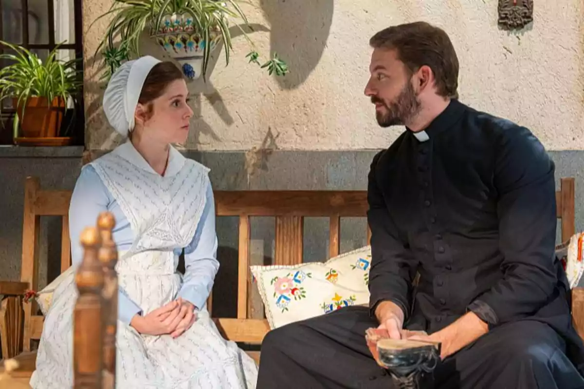 María Fernández vestida d'època i el pare Samuel amb sotana conversen asseguts a un banc de fusta en un ambient rústic amb plantes decoratives. María Fernández vestida d'època i el pare Samuel amb sotana conversen asseguts a un banc de fusta en un ambient rústic amb plantes decoratives.