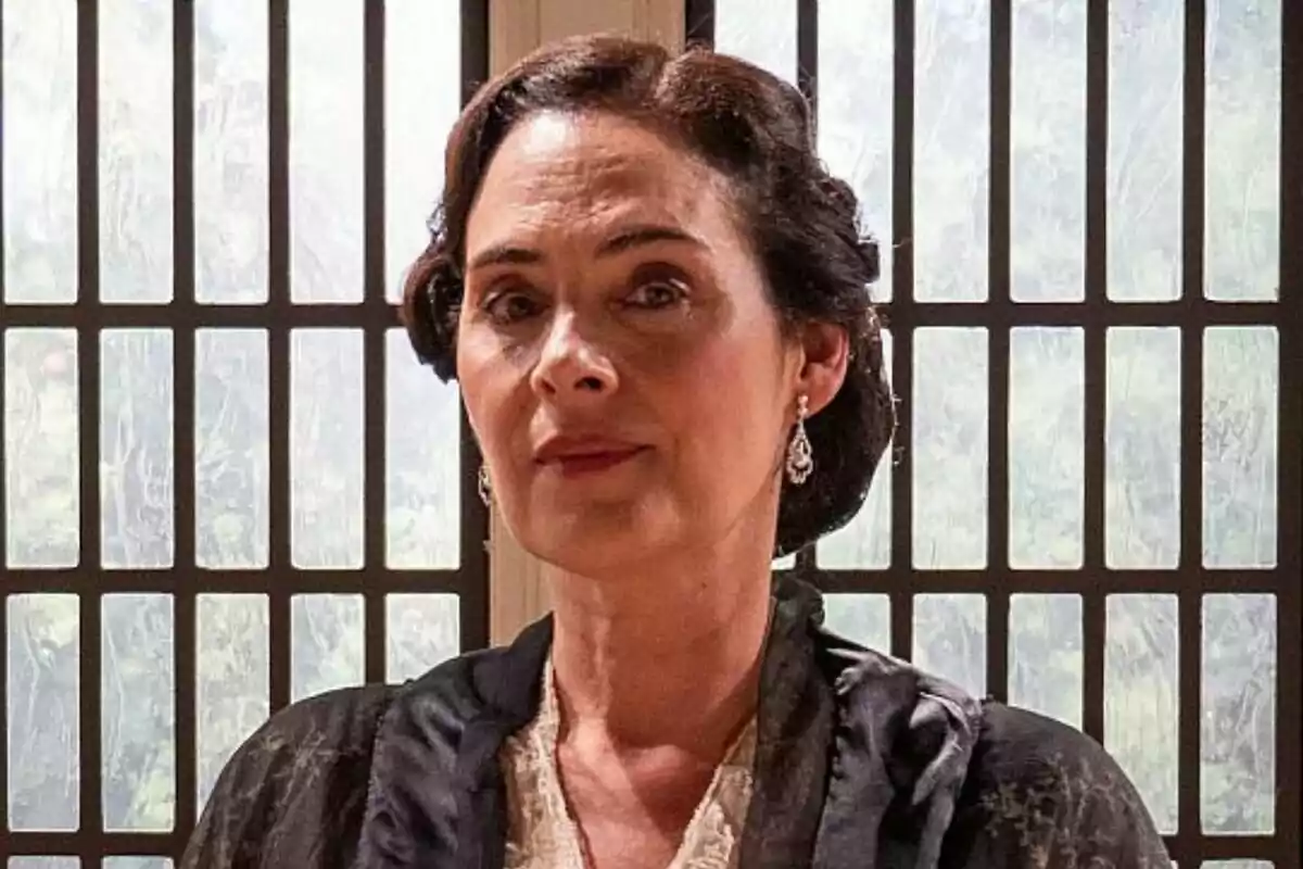 Rocío Muñoz con cabello oscuro recogido y pendientes elegantes está de pie frente a una ventana con rejas.