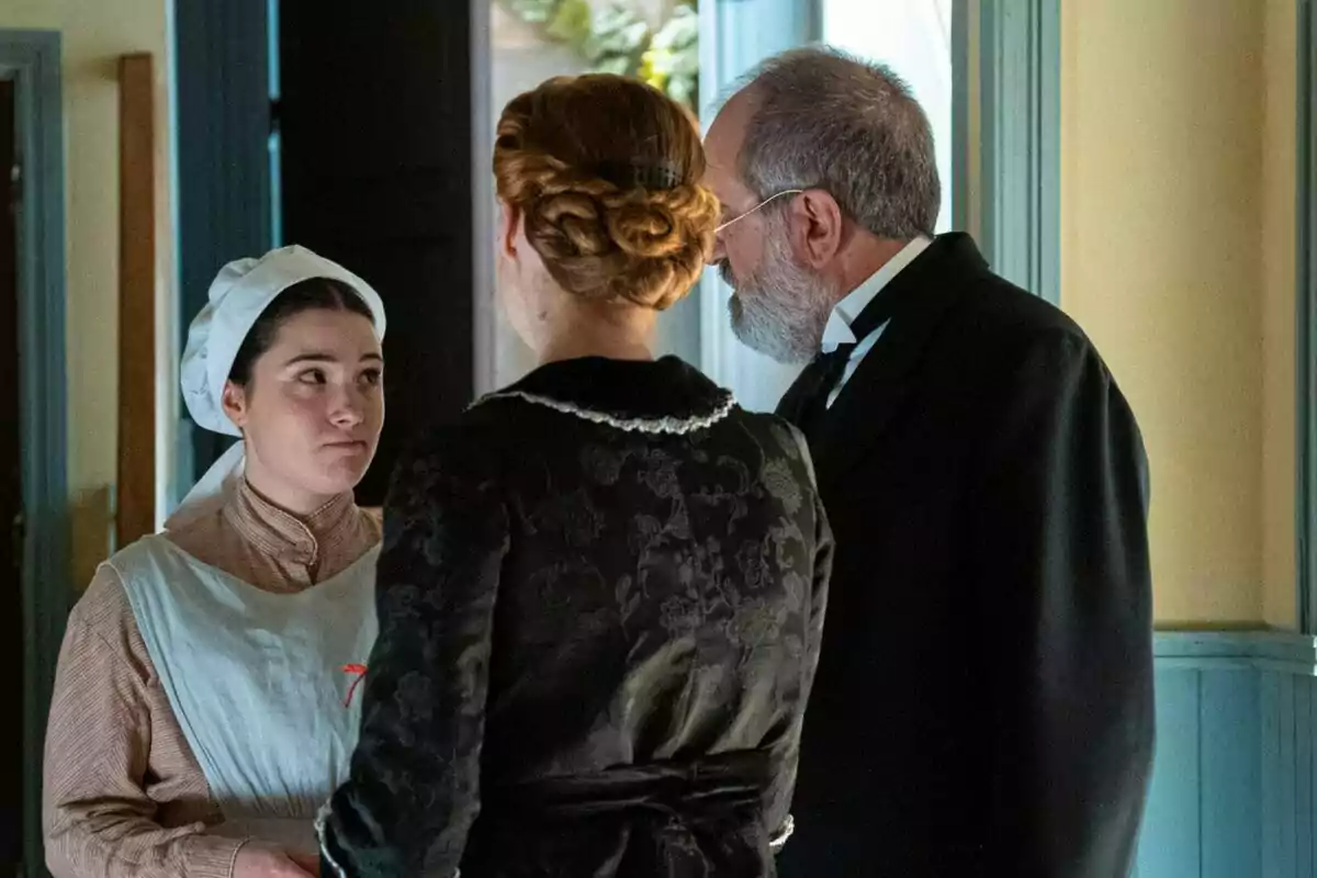 Virtudes vestida de sirvienta conversa con una mujer y un hombre mayores en una habitación iluminada. Captura de 'La Promesa' Virtudes vestida de sirvienta conversa con una mujer y un hombre mayores en una habitación iluminada. Captura de 'La Promesa'