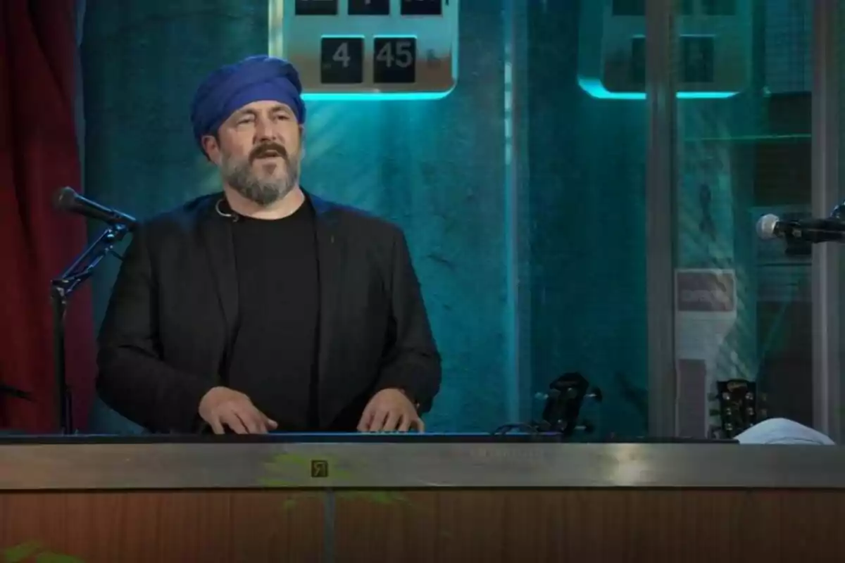 Ricardo Castella con barba y un turbante azul está de pie detrás de un teclado en un escenario iluminado con luces azules en La Revuelta. Ricardo Castella con barba y un turbante azul está de pie detrás de un teclado en un escenario iluminado con luces azules en La Revuelta.