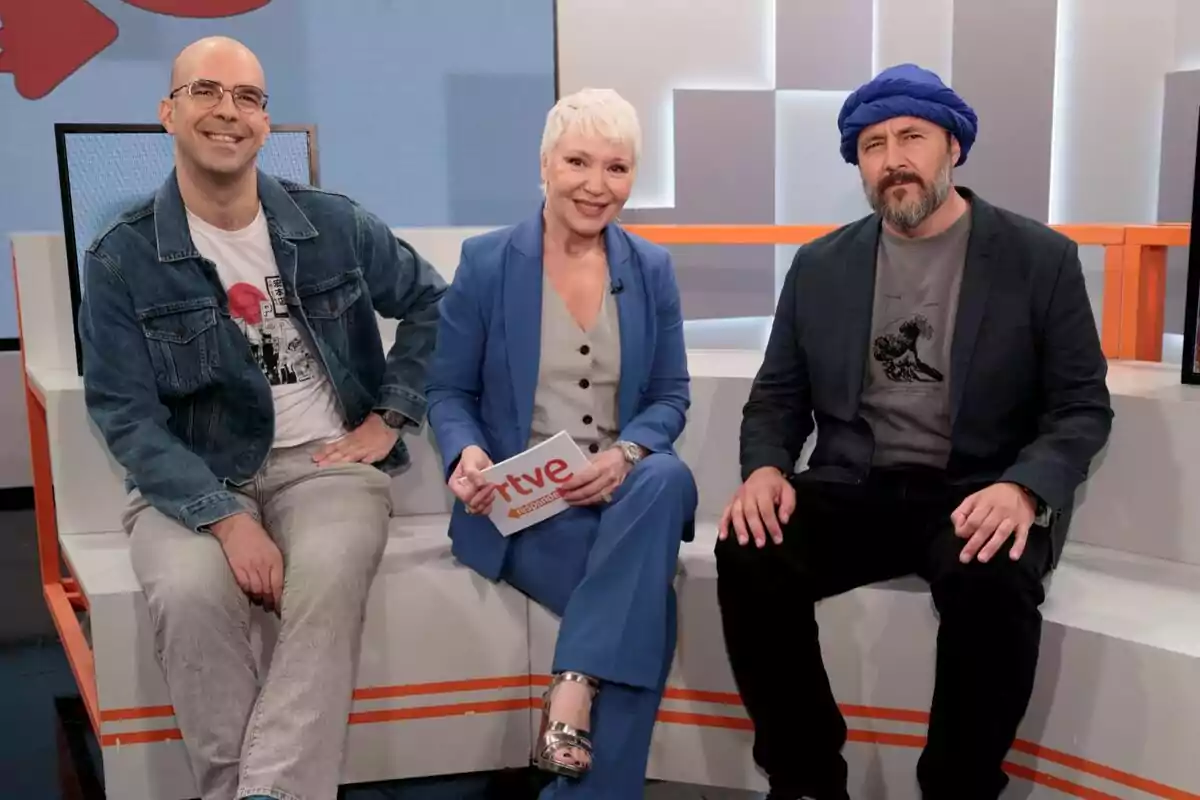Agustín Alonso, Rosa María Molló y Ricardo Castella sentados en el plató de RTVE Responde, una mujer en el centro sosteniendo una tarjeta con el logo de Agustín Alonso, Rosa María Molló y Ricardo Castella sentados en el plató de RTVE Responde, una mujer en el centro sosteniendo una tarjeta con el logo de