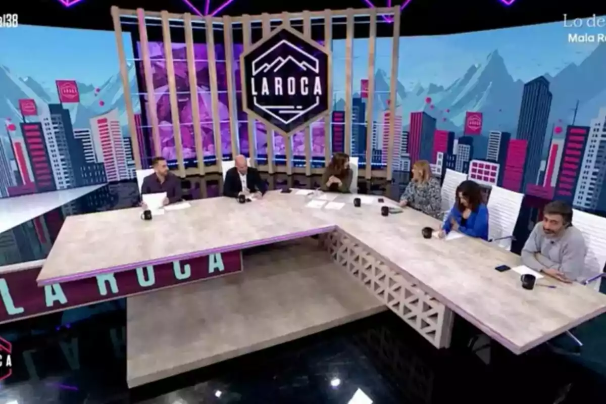 Un grup de persones està assegut al voltant d'una taula en un plató de televisió amb un fons que mostra un paisatge urbà i muntanyes, i un logotip que diu Un grup de persones està assegut al voltant d'una taula en un plató de televisió amb un fons que mostra un paisatge urbà i muntanyes, i un logotip que diu