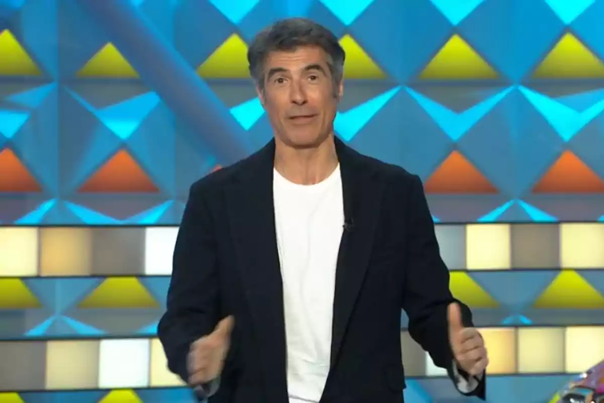 Jorge Fernández con chaqueta negra y camiseta blanca está de pie frente a un fondo colorido con patrones geométricos. Captura de 'La Ruleta de la Suerte'