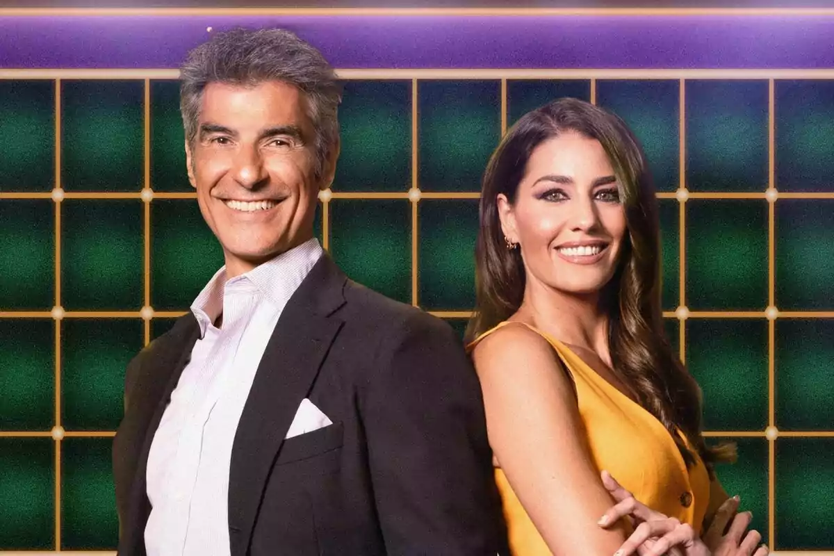 Jorge Fernández y Laura Moure sonrientes posan de pie, una al lado de la otra, con un fondo de cuadrícula verde y púrpura en La Ruleta de la Suerte. Jorge Fernández y Laura Moure sonrientes posan de pie, una al lado de la otra, con un fondo de cuadrícula verde y púrpura en La Ruleta de la Suerte.