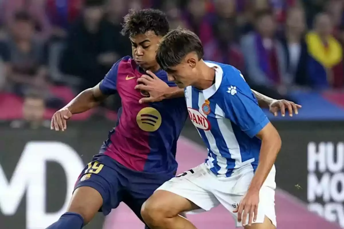 Lamine Yamal i un altre jugador de futbol competeixen per la pilota en un partit, un vesteix un uniforme blau i grana del Barça i l'altre un uniforme blau i blanc de l'Espanyol.