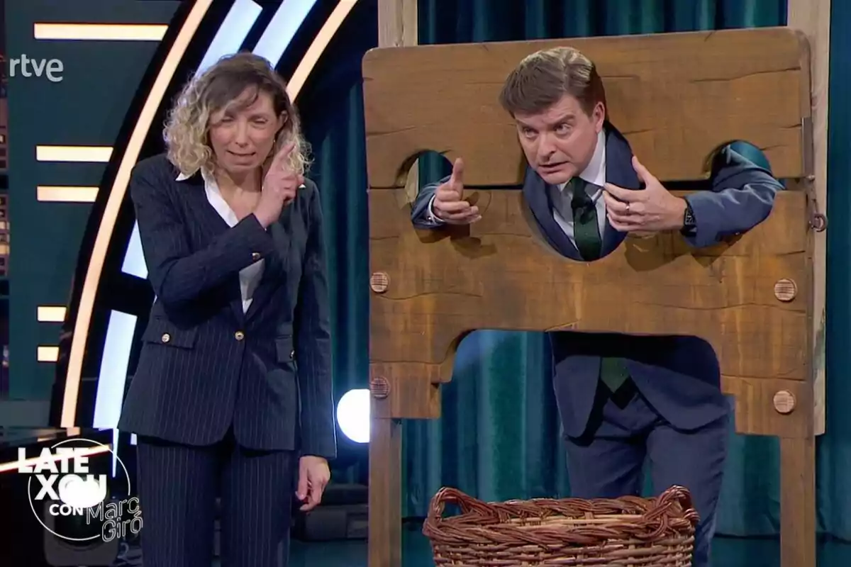 Una dona amb vestit fosc està dreta al costat de Marc Giró en un cep de fusta a 'Late Xou amb Marc Giró'. Una dona amb vestit fosc està dreta al costat de Marc Giró en un cep de fusta a 'Late Xou amb Marc Giró'.