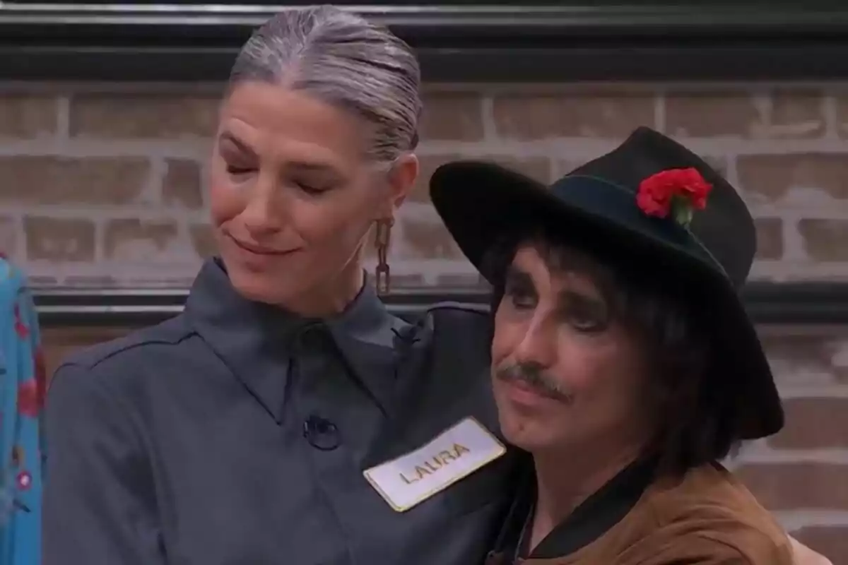 Laura Sánchez con cabello canoso y peinado hacia atrás, usando un uniforme gris con una etiqueta que dice "Laura", está abrazando a Canco Rodríguez que lleva un sombrero negro adornado con una flor roja en Maestros de la Costura Celebrity.