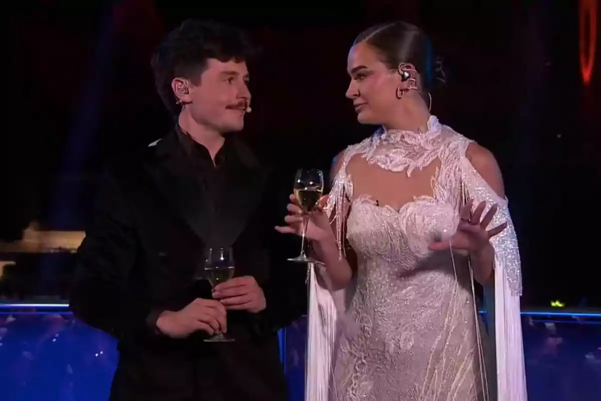 Miki Núñez y Laura Escanes elegantemente vestidas sostienen copas de vino mientras se miran en un escenario iluminado en las Campanadas de TV3.
