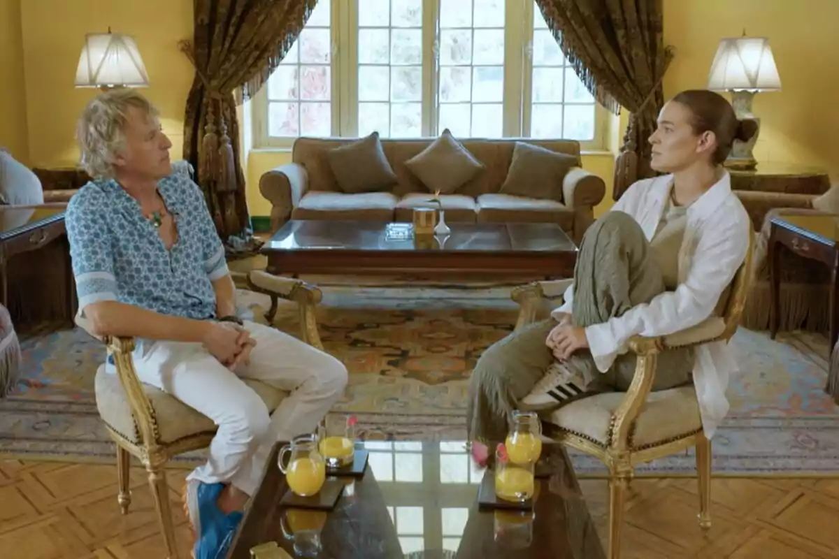 Laura Escanes y Jesús Calleja sentadas en una sala elegante con cortinas oscuras y muebles clásicos, conversando frente a una mesa de centro con vasos de jugo de naranja en Universo Calleja. Laura Escanes y Jesús Calleja sentadas en una sala elegante con cortinas oscuras y muebles clásicos, conversando frente a una mesa de centro con vasos de jugo de naranja en Universo Calleja.