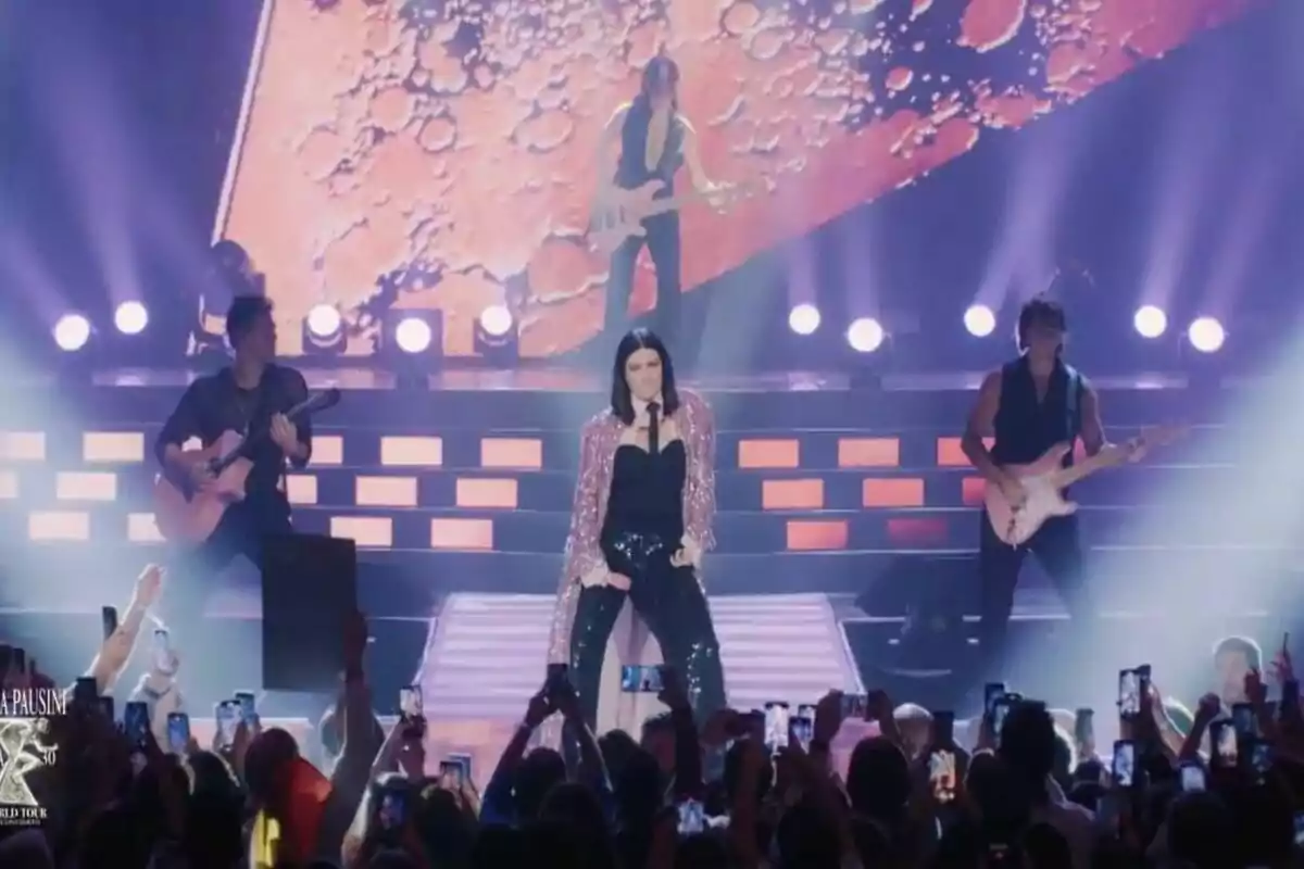 Laura Pausini en el escenario con una chaqueta brillante y pantalones negros, acompañada por músicos con guitarras, mientras el público levanta sus teléfonos para grabar el espectáculo. Laura Pausini en el escenario con una chaqueta brillante y pantalones negros, acompañada por músicos con guitarras, mientras el público levanta sus teléfonos para grabar el espectáculo.