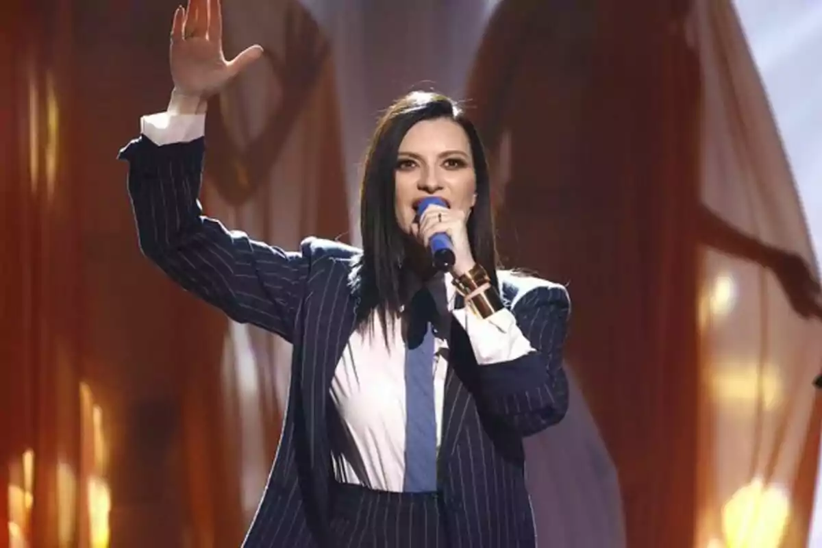 Laura Pausini con traje a rayas canta en un escenario iluminado mientras levanta una mano en La Voz.