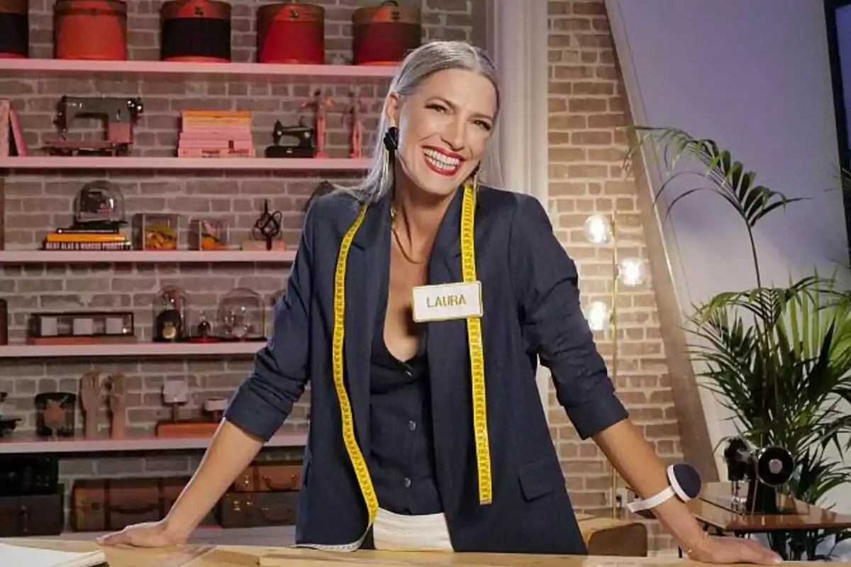 Laura Sánchez sonriente con cabello canoso y cinta métrica amarilla alrededor del cuello, de pie en un taller de costura con estantes llenos de objetos detrás en Maestros de la Costura Celebrity. Laura Sánchez sonriente con cabello canoso y cinta métrica amarilla alrededor del cuello, de pie en un taller de costura con estantes llenos de objetos detrás en Maestros de la Costura Celebrity.