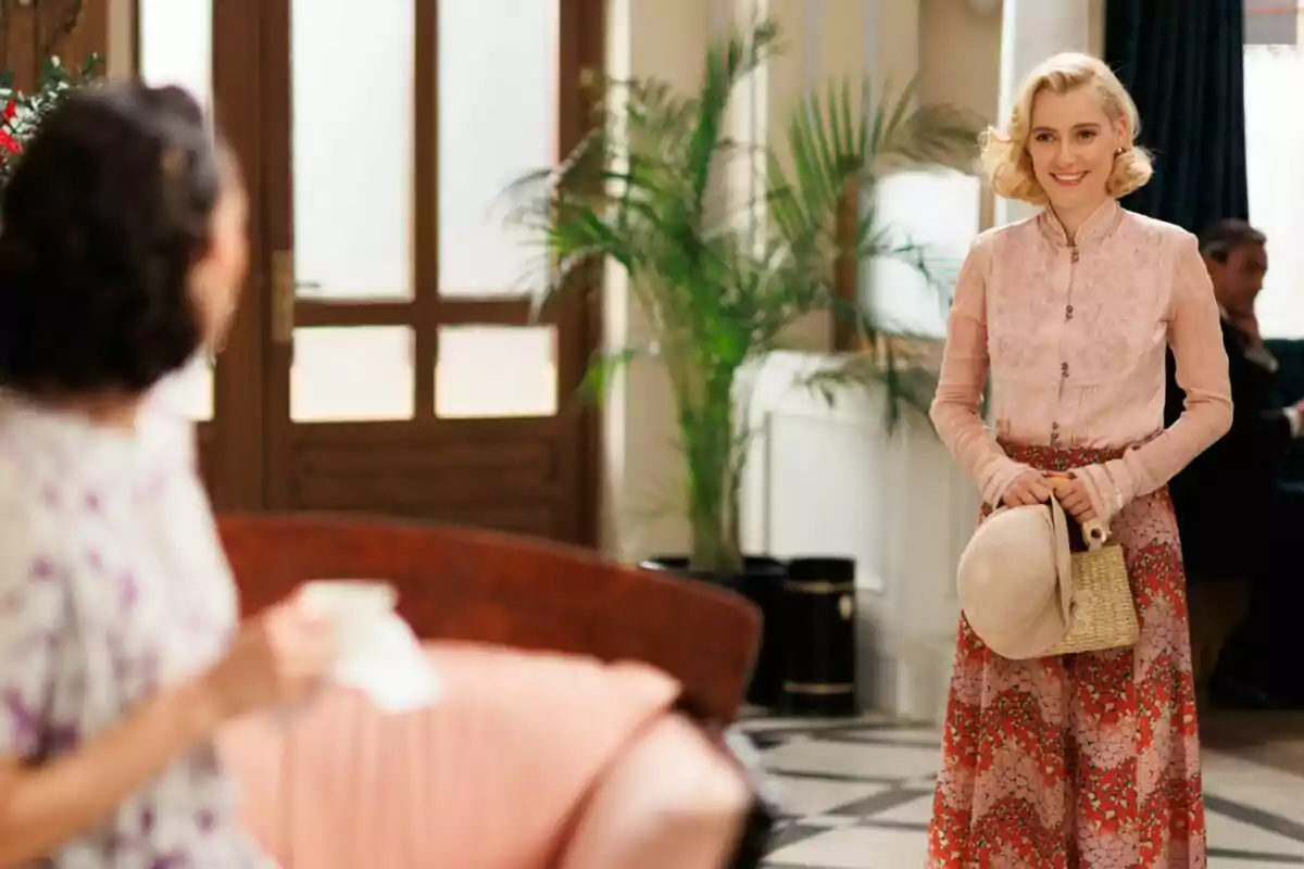 Laurita rubia con una blusa rosa y una falda larga estampada sostiene un sombrero y una bolsa de mimbre mientras sonríe en un interior elegante con plantas y muebles clásicos en La Moderna. Laurita rubia con una blusa rosa y una falda larga estampada sostiene un sombrero y una bolsa de mimbre mientras sonríe en un interior elegante con plantas y muebles clásicos en La Moderna.