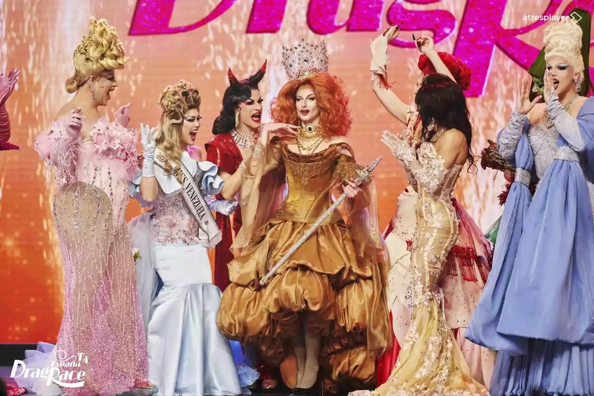 Le Cocó juntament amb un grup de persones en elaborats vestits de gala celebra en un escenari amb fons brillant a Drag Race España.