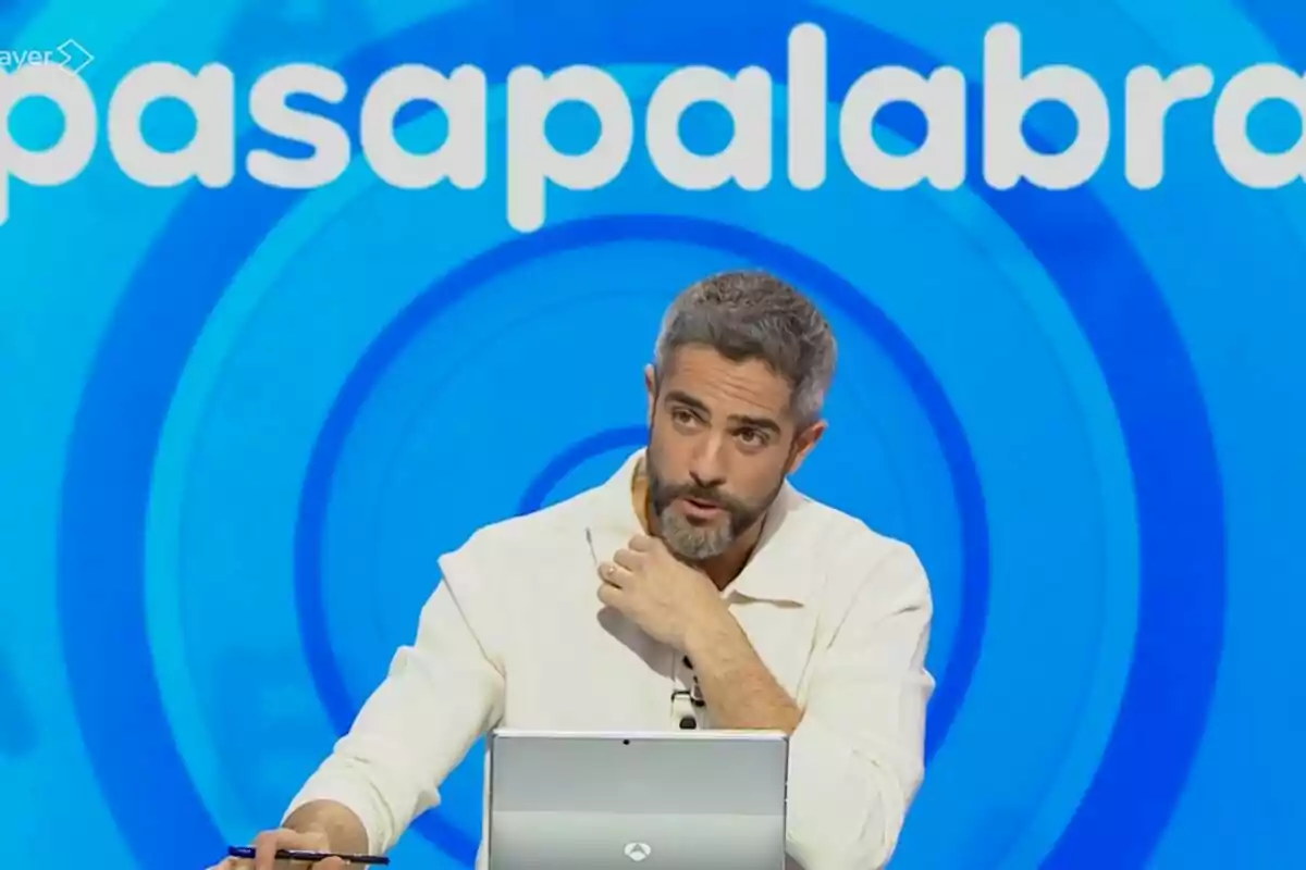 Roberto Leal con barba y cabello canoso está sentado frente a una computadora portátil en un set de televisión con un fondo azul que tiene el texto Roberto Leal con barba y cabello canoso está sentado frente a una computadora portátil en un set de televisión con un fondo azul que tiene el texto
