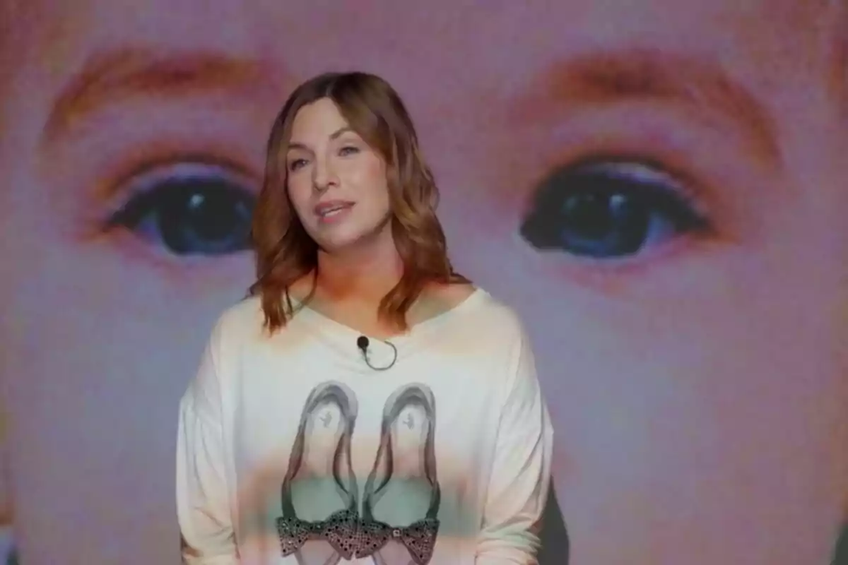 Pla mitjà de Leire Martínez vestint una samarreta amb un disseny de sabates està dret davant d'una imatge ampliada d'uns ulls. Pla mitjà de Leire Martínez vestint una samarreta amb un disseny de sabates està dret davant d'una imatge ampliada d'uns ulls.