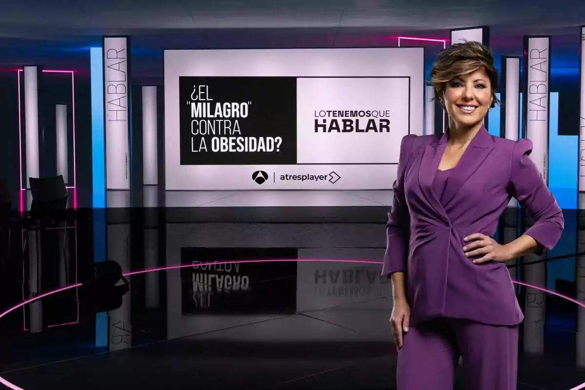 Sonsoles Ónega somrient amb un vestit morat està en un set de televisió modern amb llums de neó i pantalles que mostren el text Sonsoles Ónega somrient amb un vestit morat està en un set de televisió modern amb llums de neó i pantalles que mostren el text