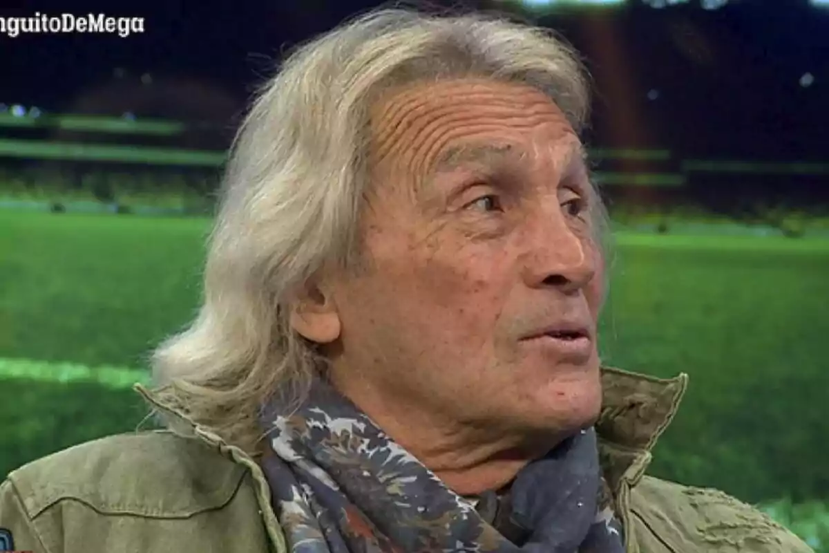 Loco Gatti de cabell canós i llarg, amb una jaqueta verda i una bufanda estampada, està en un entorn que sembla un estudi de televisió amb un fons que simula un camp de futbol a El Chiringuito de Jugones.