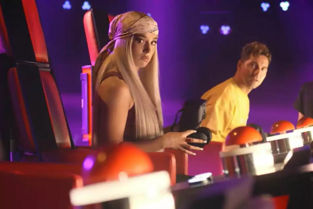 David Bisbal y Lola Índigo en sillas rojas de La Voz Kids, una mujer con pañuelo en la cabeza y un hombre con camiseta amarilla, ambos mirando hacia adelante.