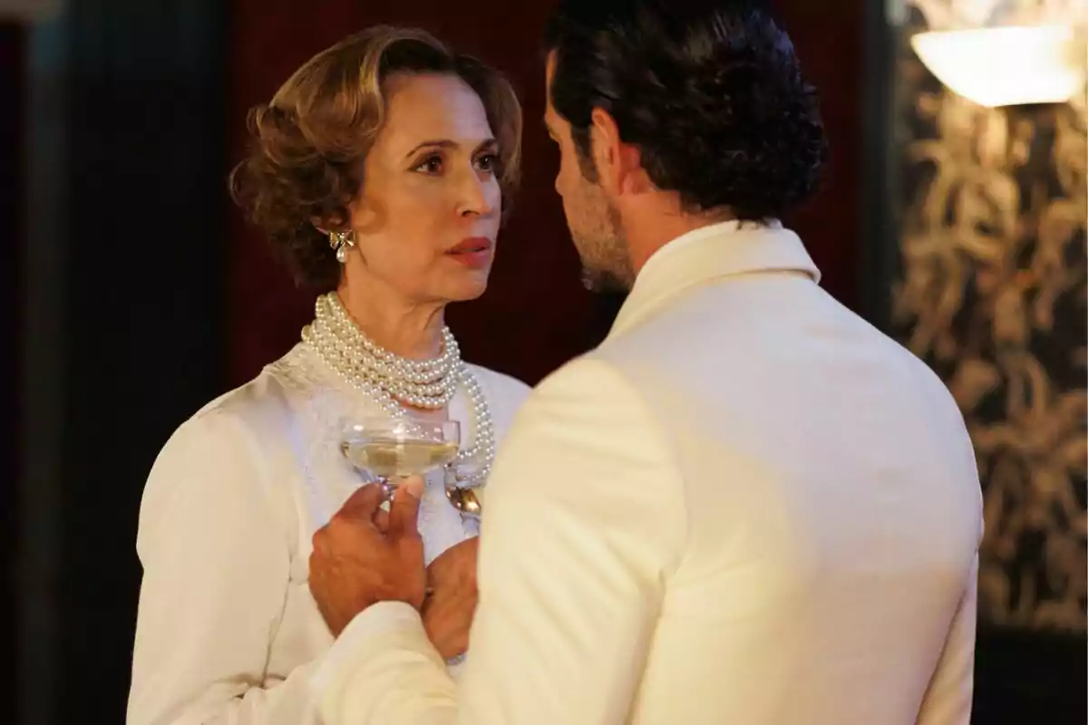 Lola Marceli y Miguel Ángel Muñoz elegantemente vestidos se miran fijamente mientras ella sostiene una copa en La Moderna. Lola Marceli y Miguel Ángel Muñoz elegantemente vestidos se miran fijamente mientras ella sostiene una copa en La Moderna.